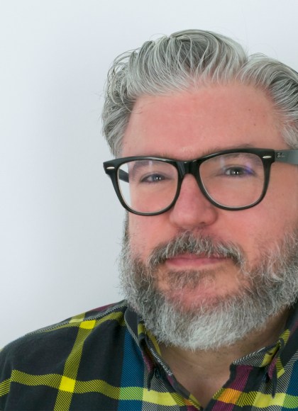 Photo of author Alonso Duralde. The author has salt and pepper hair, mustache, and beard, and wears black glasses. He is looking at the camera in a candid shot.
