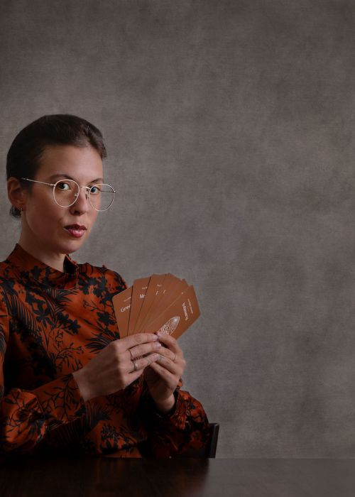 Photo of author Maude White. The author wears glasses and holds cards from the Pleasure Alchemy deck. She has a mysterious look on her face.