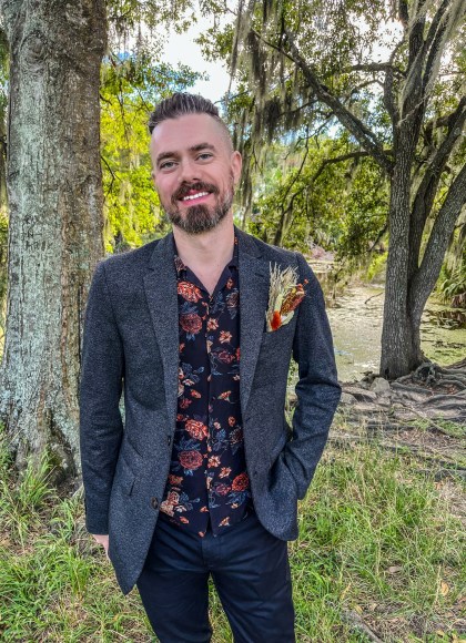 Photo of author Alexander Schneider. The author is standing in a clearing among trees, wearing a casual three-piece suit with a boutonnière.
