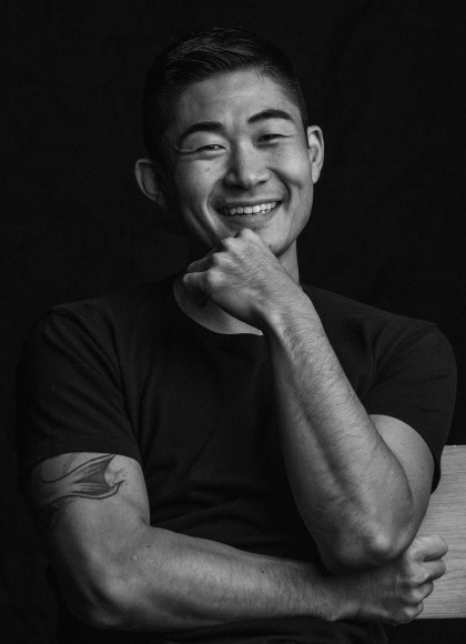 Black and white photo of artist Yoshino from the waist up. The artist is wearing a black shirt, and has one arm crossed in front with the other elbow sitting on that arm and raised up to his chin. He is smiling into the camera.